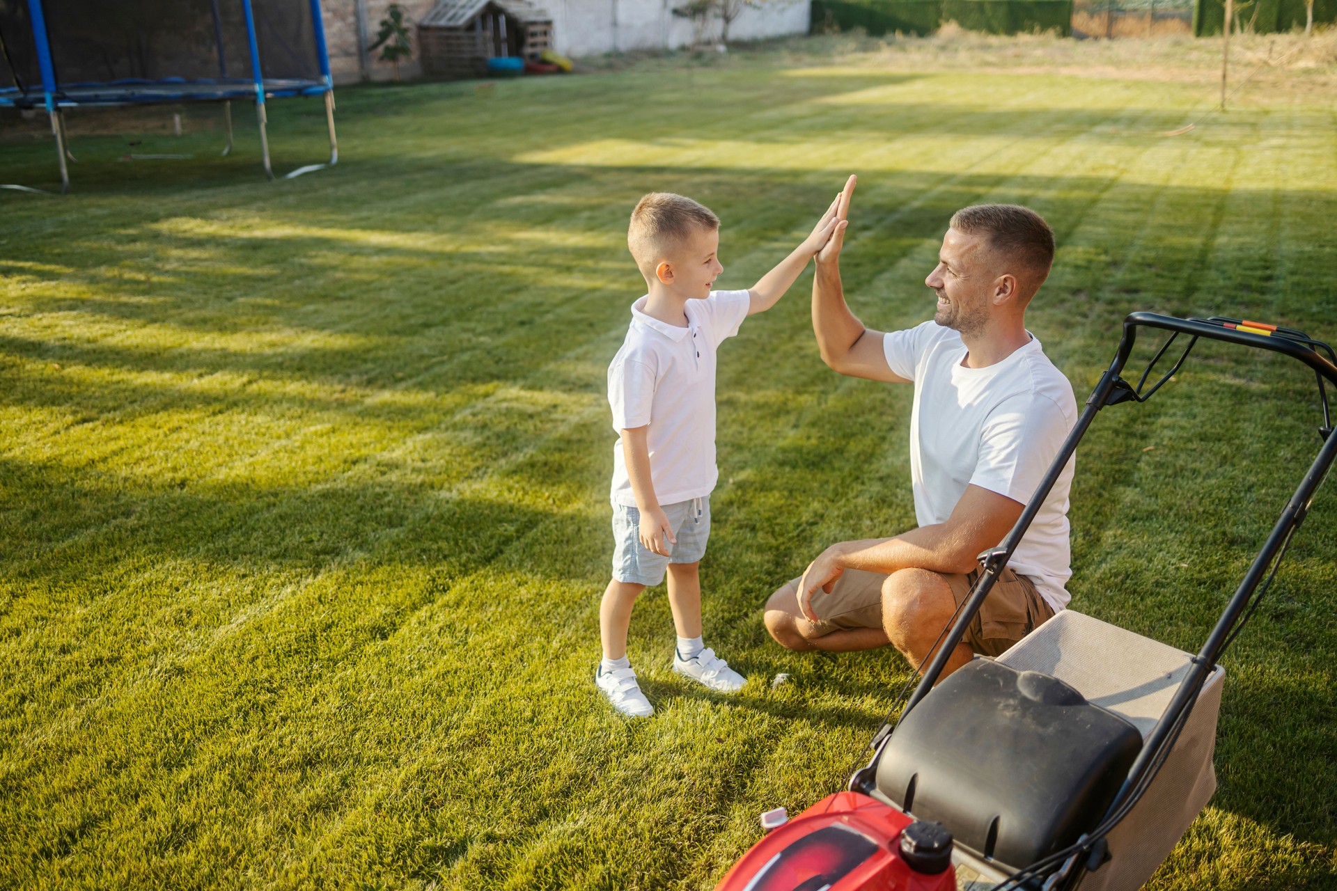 Teamwork. Father and his little son giving high five to each other for finishing their gardening chore with grass mower.